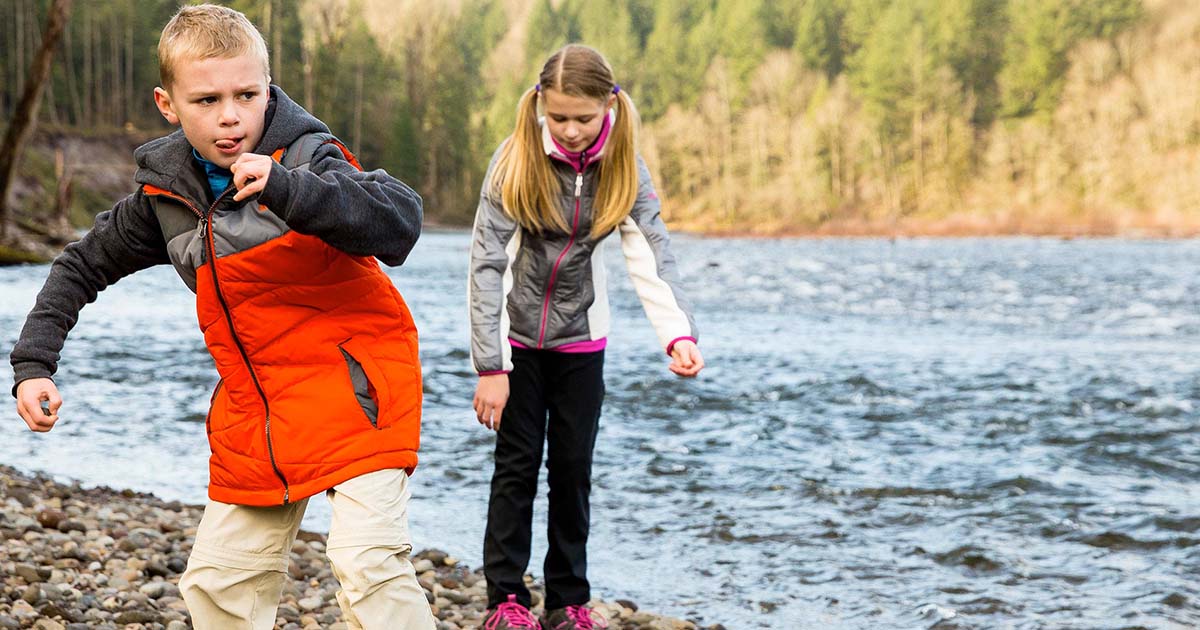 A boy winds up to throw a rock while a girl inspects rocks beside a rushing river